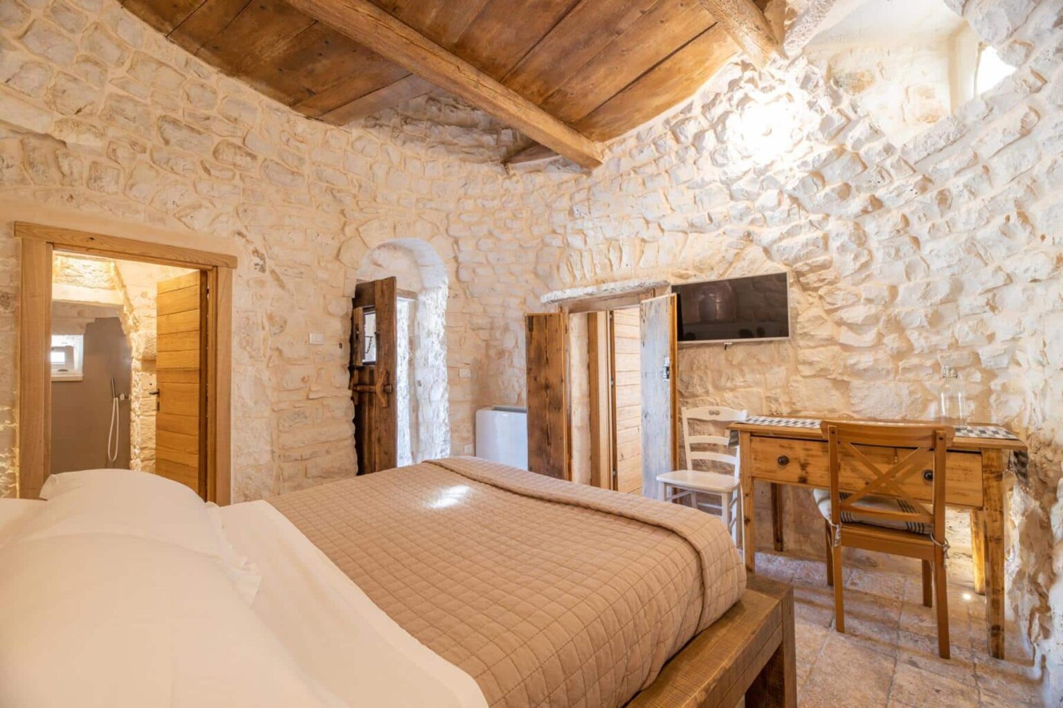 Cozy stone bedroom featuring a comfortable bed, wooden ceiling beams, and rustic decor. The room includes a desk with chairs, a flat-screen TV, and an en-suite bathroom visible through an open door. Ideal for a relaxing getaway. | Only on SopranoVillas