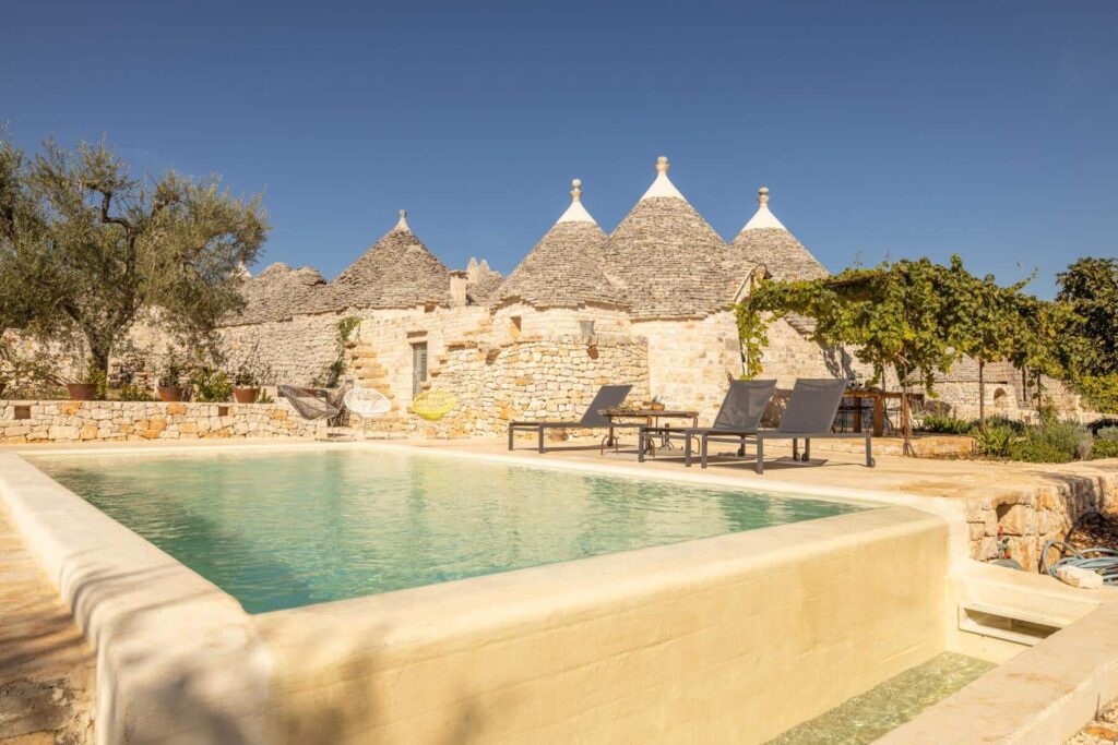 Tranquil swimming pool surrounded by stone trulli houses in a sunny landscape, ideal for a relaxing getaway in Puglia, Italy. | Only on SopranoVillas