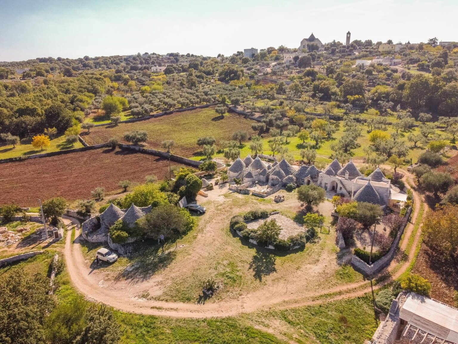 Aerial view of traditional trulli houses surrounded by olive groves and farmland in the countryside, showcasing rural architecture and natural landscapes. | Only on SopranoVillas