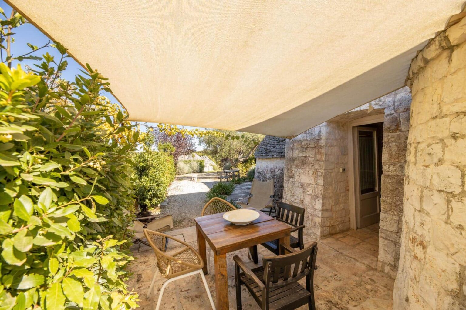 Cozy outdoor dining area featuring a wooden table and chairs shaded by a fabric canopy, surrounded by lush greenery and stone walls, ideal for relaxation and meals in nature. | Only on SopranoVillas