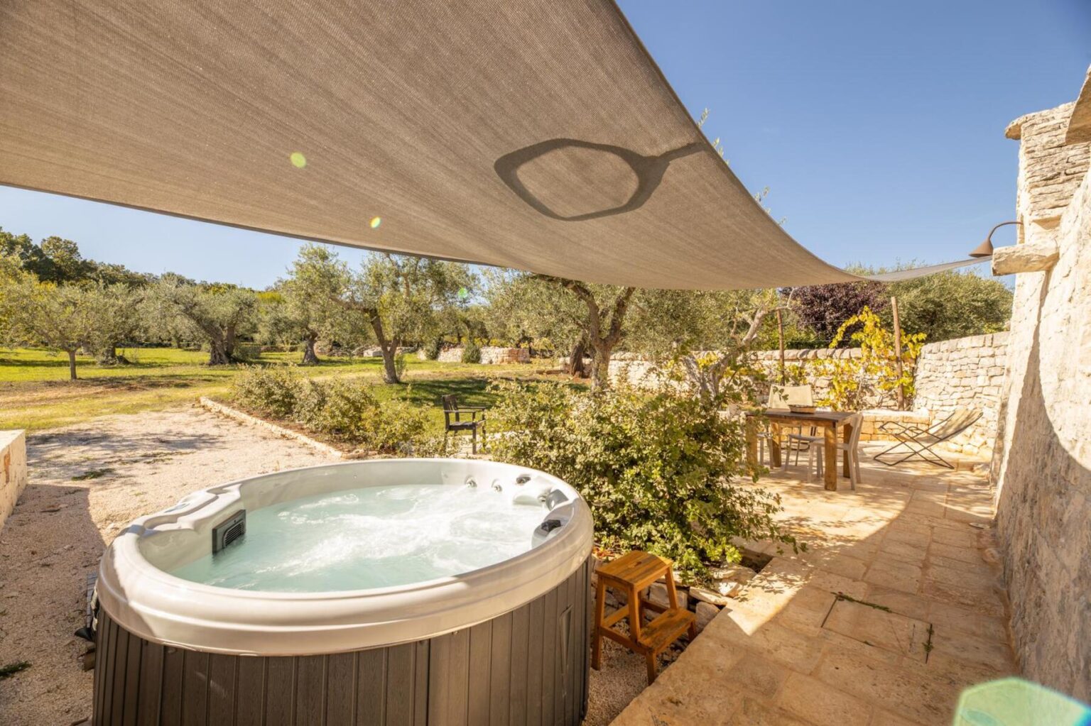 Relaxing outdoor hot tub under a shaded canopy in a serene garden setting with olive trees and rustic stone walls. Perfect for a tranquil getaway. | Only on SopranoVillas