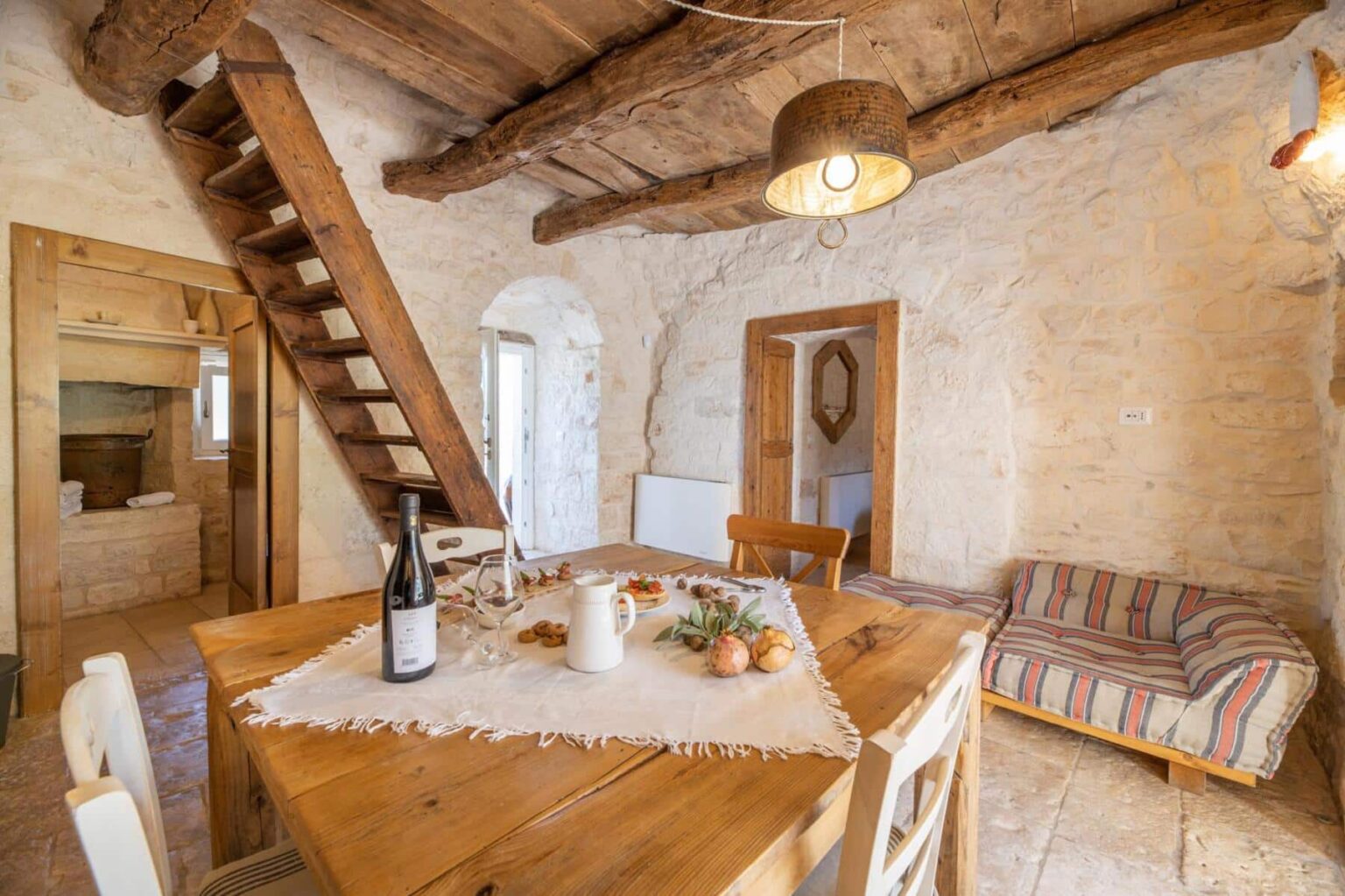 Cozy rustic dining area with wooden table, natural light, and a staircase leading to an upper level, featuring a wine bottle and decorative items. Stone walls and wooden beams create a warm, inviting atmosphere. | Only on SopranoVillas