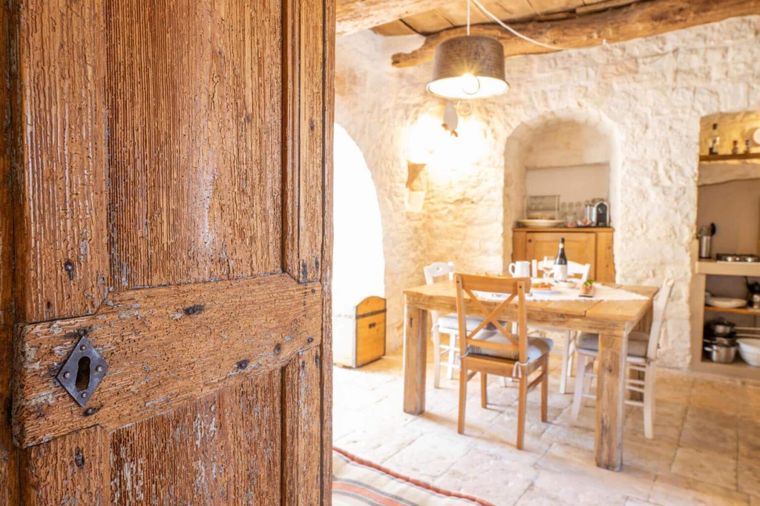 Rustic wooden door partially open, revealing a cozy kitchen and dining area with a wooden table, chairs, and soft lighting. Natural stone walls and a warm ambiance create an inviting atmosphere. | Only on SopranoVillas