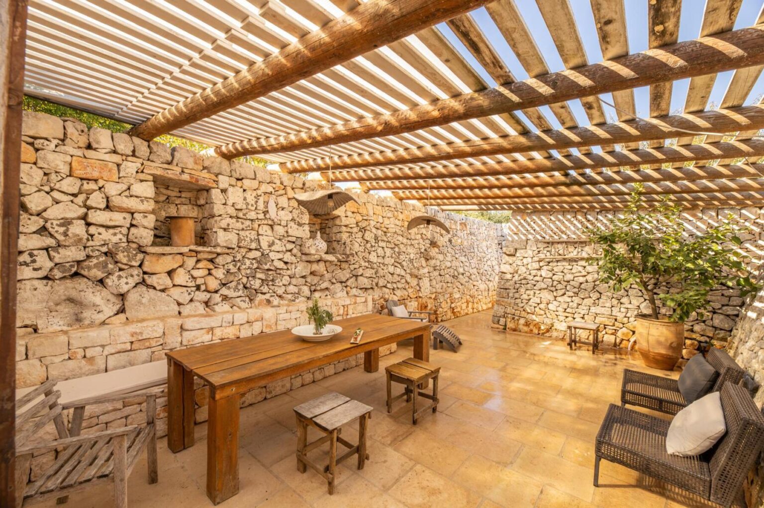 Cozy outdoor dining area featuring a wooden table and chairs, surrounded by rustic stone walls and shaded by a slatted pergola, ideal for al fresco meals. | Only on SopranoVillas