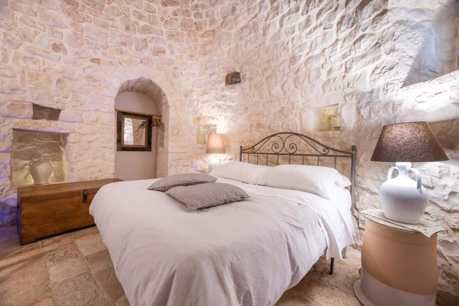 Cozy bedroom featuring a stone wall, a comfortable double bed with decorative pillows, a wooden chest, and a stylish lamp on a unique side table, creating a warm and inviting atmosphere. | Only on SopranoVillas