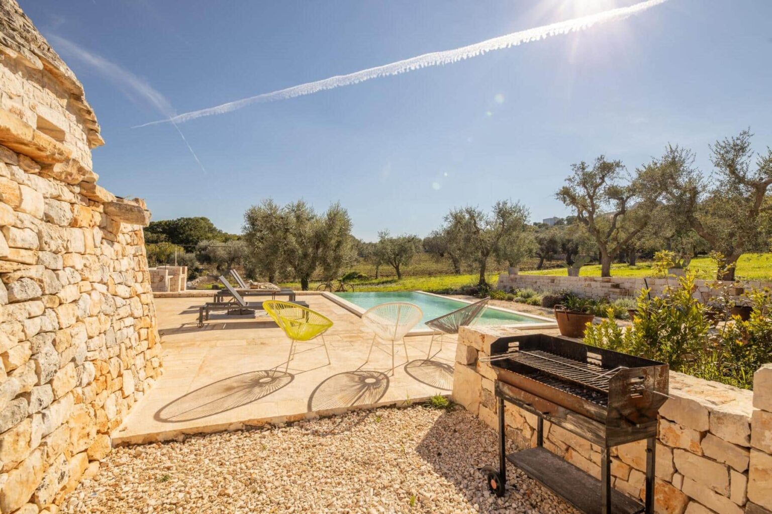 Scenic view of a rustic stone patio featuring a swimming pool, lounge chairs, and a barbecue grill, surrounded by olive trees and lush greenery under a clear blue sky. | Only on SopranoVillas