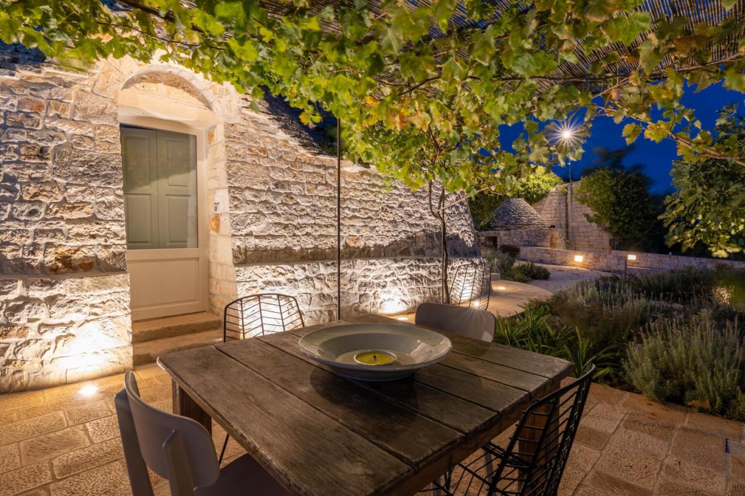 Cozy outdoor dining area illuminated at night, featuring a wooden table, modern chairs, and a stone wall backdrop surrounded by lush greenery and soft lighting. | Only on SopranoVillas