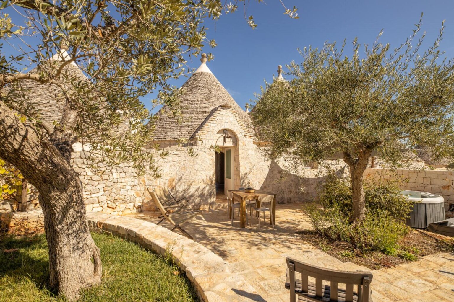 Trullo house in Apulia, Italy, featuring traditional conical roofs, surrounded by olive trees and a stone patio with outdoor seating. | Only on SopranoVillas