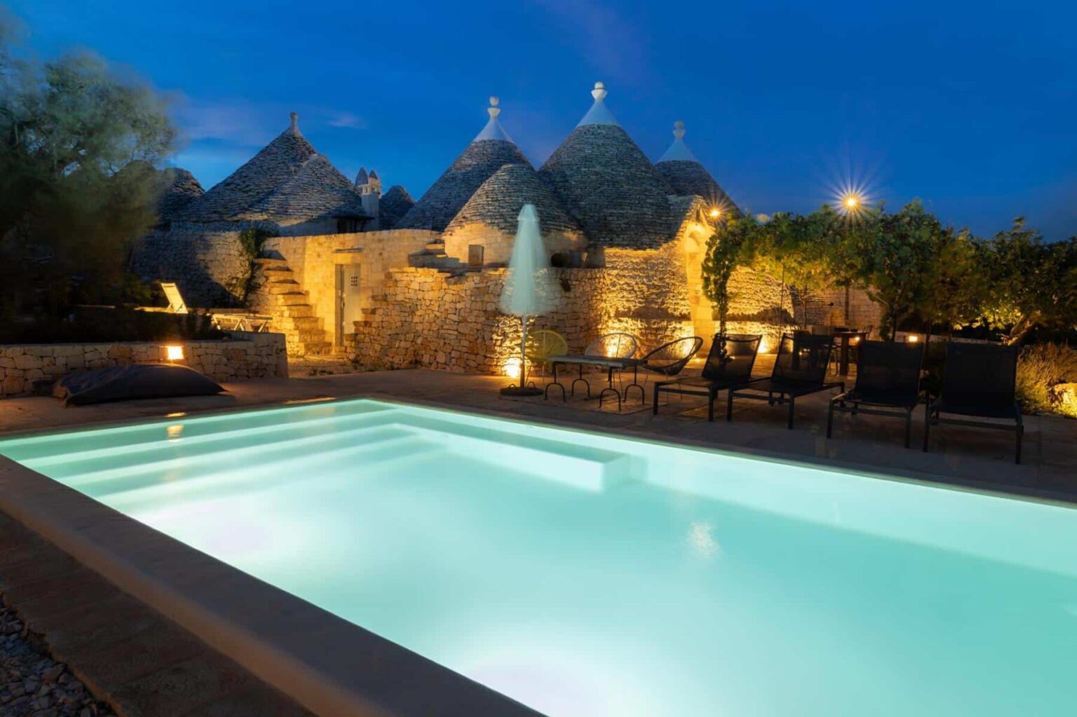 Tranquil evening scene featuring a modern swimming pool surrounded by traditional trulli houses, illuminated by soft lighting. Comfortable lounge chairs are placed nearby, enhancing the serene atmosphere of this picturesque outdoor space. | Only on SopranoVillas