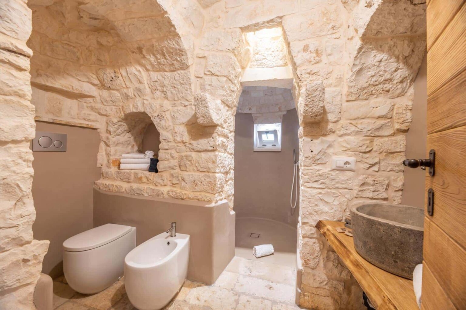 Modern bathroom featuring natural stone walls, a sleek toilet, bidet, and a walk-in shower. The design includes wooden accents, neatly stacked towels, and a small window for natural light, creating a stylish and inviting space. | Only on SopranoVillas