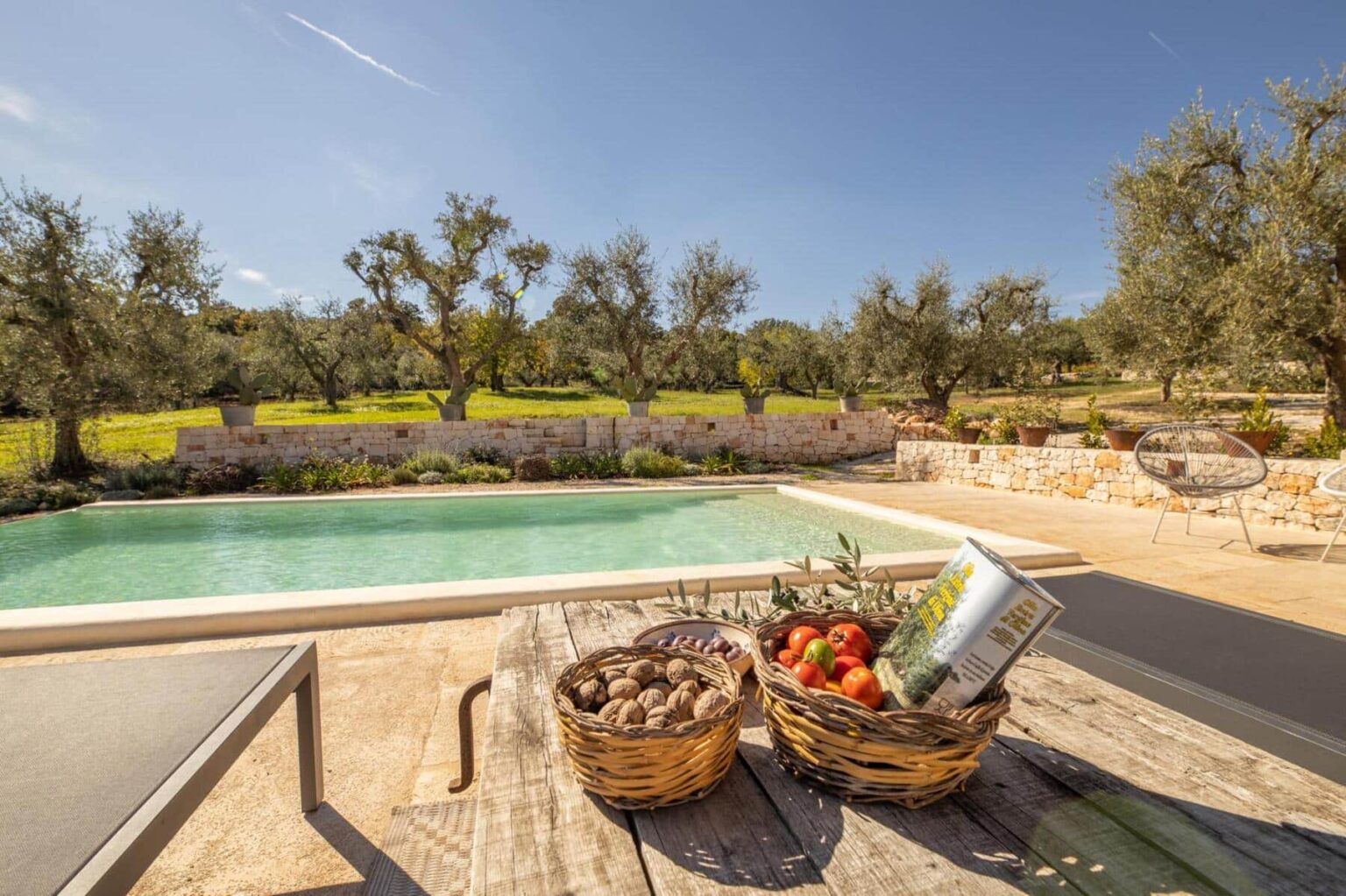 Scenic view of a tranquil poolside area surrounded by olive trees, featuring wooden tables with baskets of fresh fruits and vegetables, ideal for outdoor relaxation and dining in a sunny landscape. | Only on SopranoVillas