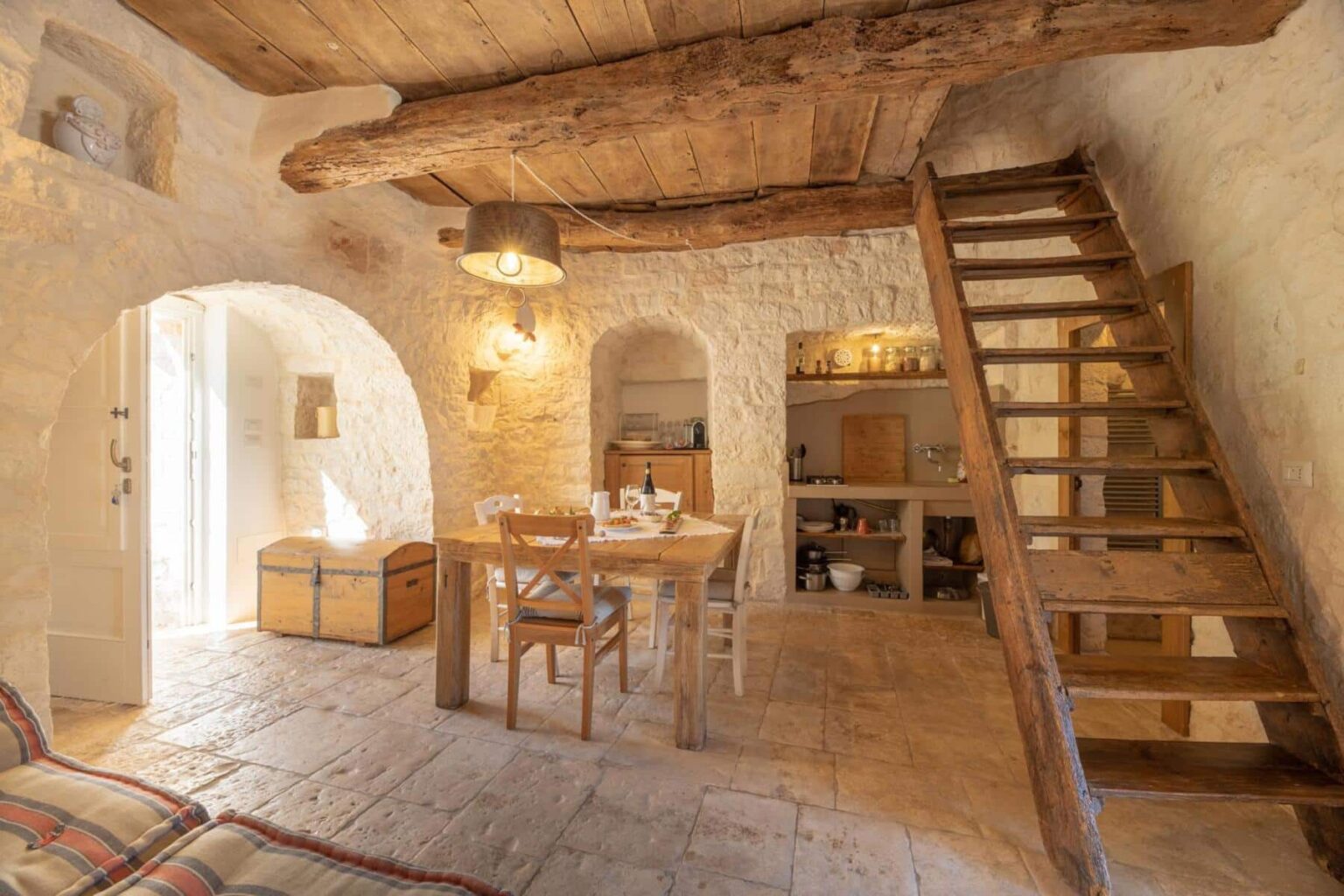 Cozy interior of a rustic home featuring exposed wooden beams, a wooden dining table set for meals, and a staircase leading to an upper level, with light-colored stone walls and warm lighting. | Only on SopranoVillas