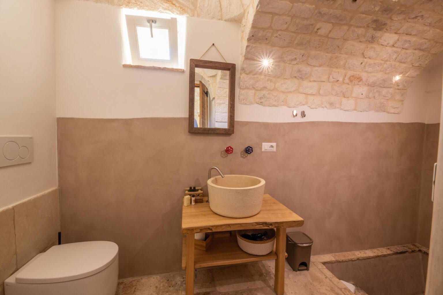 Modern bathroom interior featuring a stone arch ceiling, minimalist design, wooden vanity with a round sink, and soft neutral tones. Includes a toilet, mirror, and stylish decor elements. Ideal for showcasing contemporary bath design. | Only on SopranoVillas