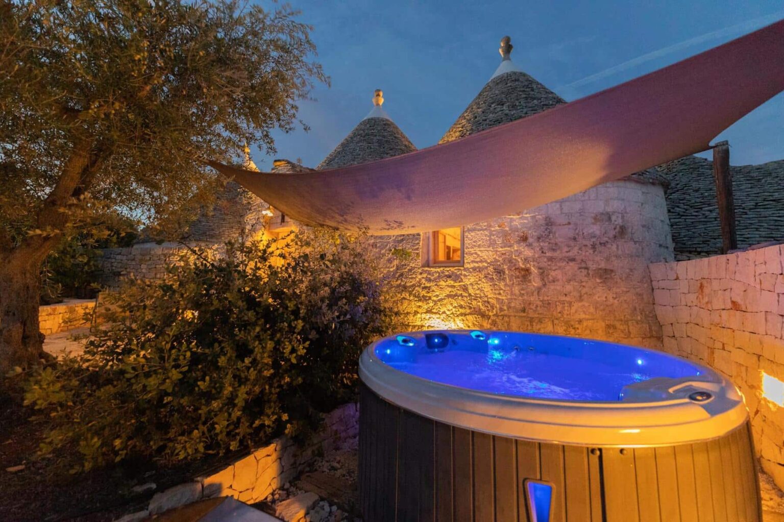 Outdoor hot tub illuminated at night, surrounded by rustic stone walls and traditional trulli structures, creating a serene and relaxing atmosphere. | Only on SopranoVillas