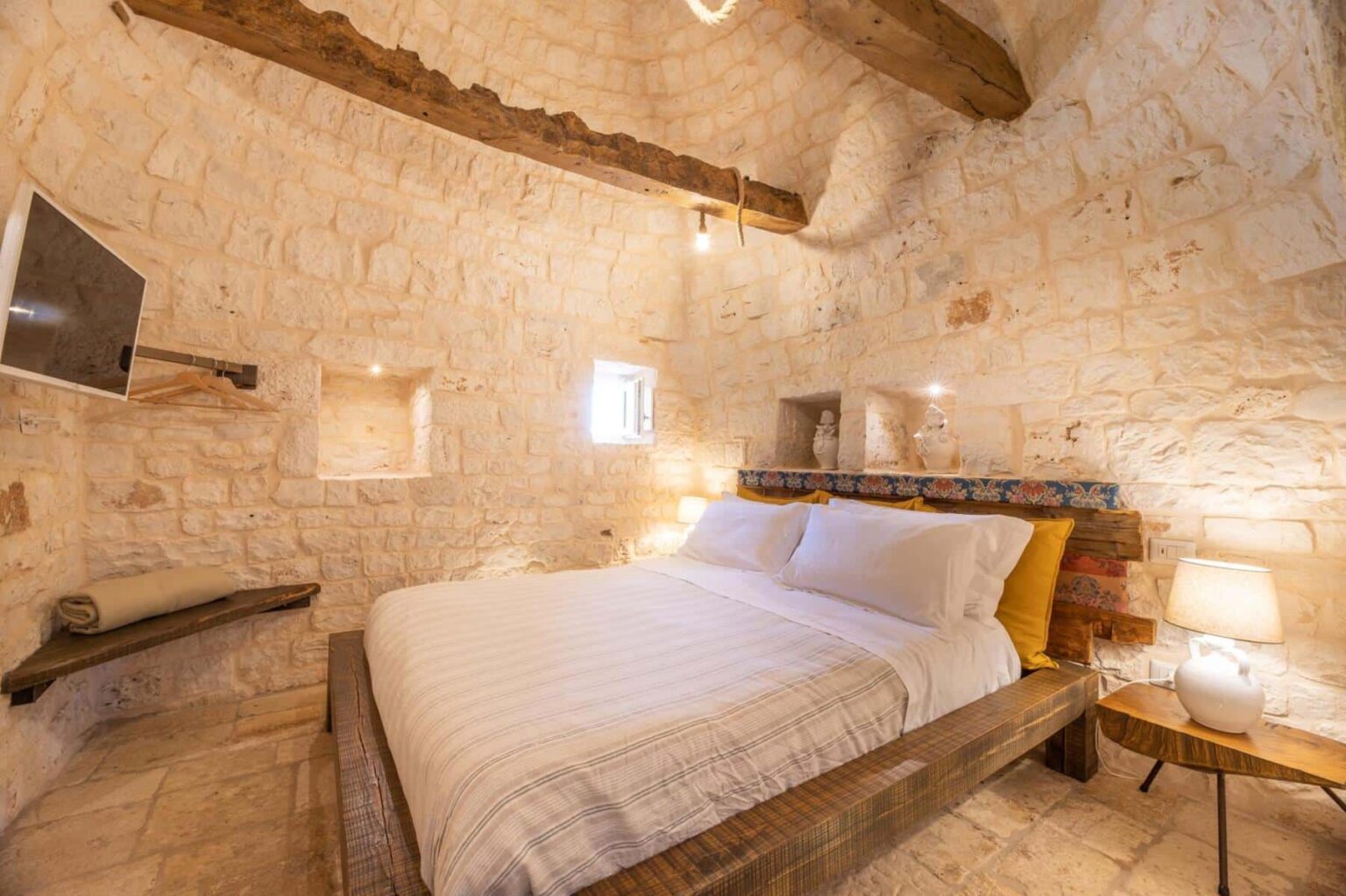 Cozy bedroom featuring a wooden bed with white linens, stone walls, and rustic wooden beams, complemented by soft lighting and a small TV, ideal for a relaxing stay. | Only on SopranoVillas