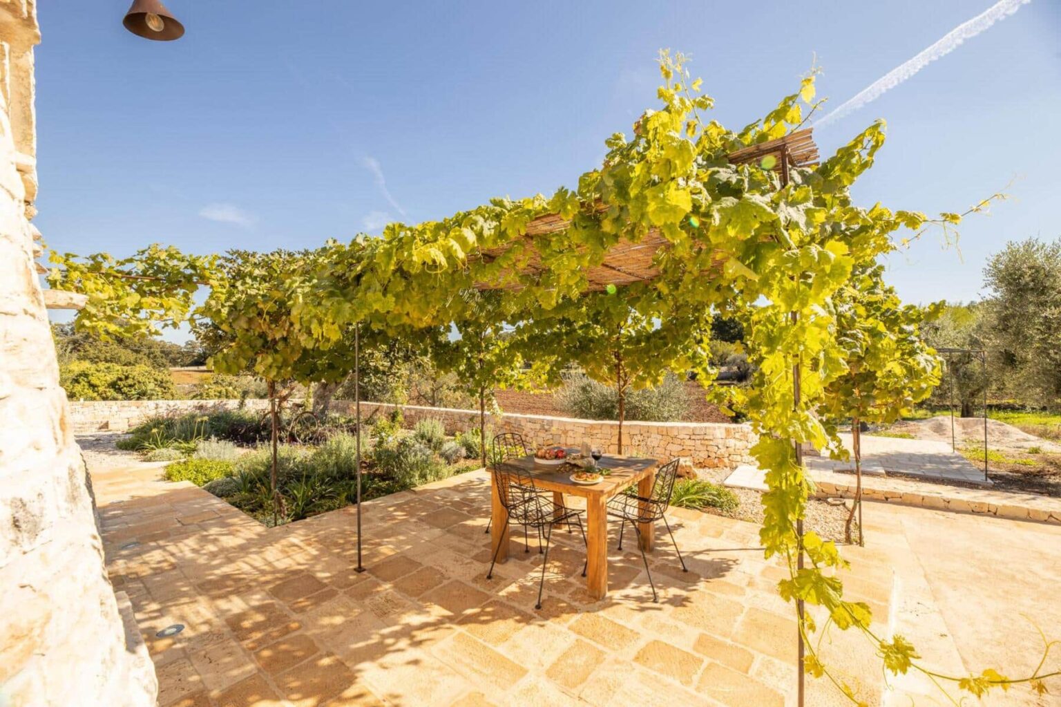 Outdoor dining area under a grapevine canopy, featuring a wooden table set with food and drinks. Surrounded by lush greenery and stone walls, the scene captures a serene countryside atmosphere with clear blue skies. Perfect for al fresco dining and relaxation. | Only on SopranoVillas
