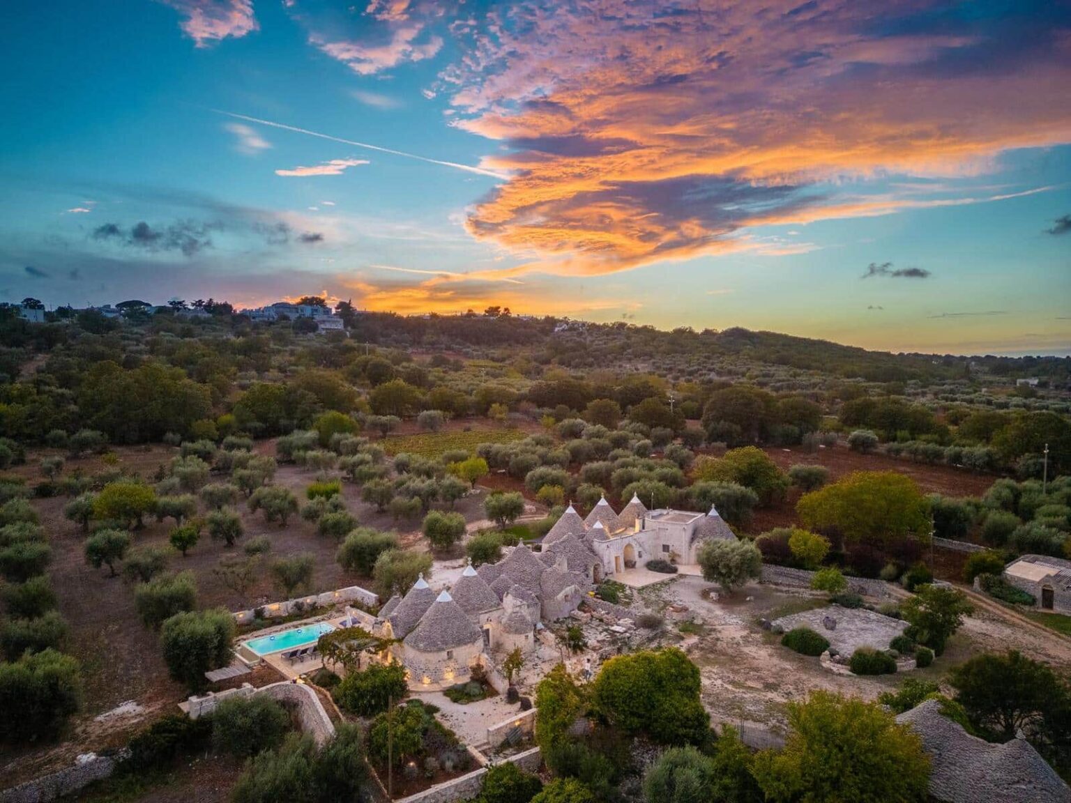 Aerial view of a charming trulli house surrounded by lush olive groves at sunset, showcasing a serene landscape with a swimming pool and vibrant sky colors. Ideal for vacation rentals in Puglia, Italy. | Only on SopranoVillas