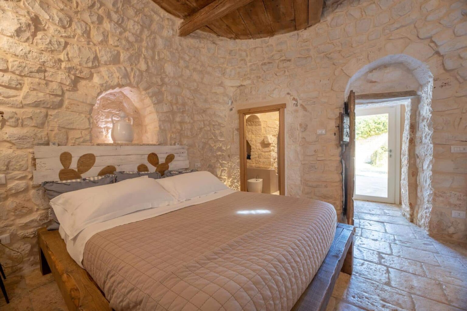 Cozy bedroom with a stone wall, featuring a wooden bed with a quilted cover and decorative pillows. The room has a rustic charm with arched doorways and warm lighting, offering a peaceful ambiance. A doorway leads to an outdoor area, enhancing the inviting atmosphere. | Only on SopranoVillas