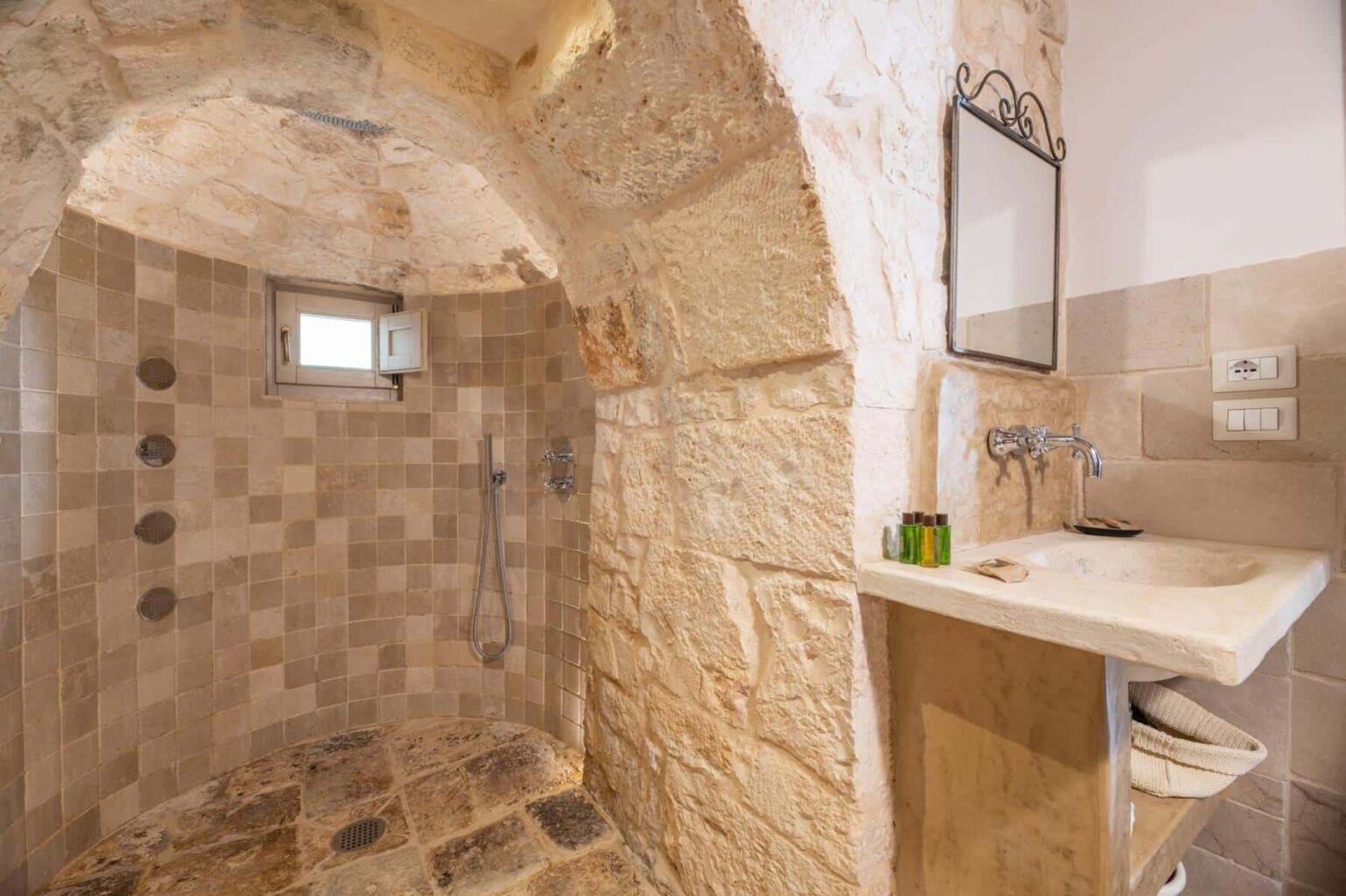 Modern bathroom with a stone shower, natural light, and a minimalist sink area featuring a mirror and decorative elements. | Only on SopranoVillas