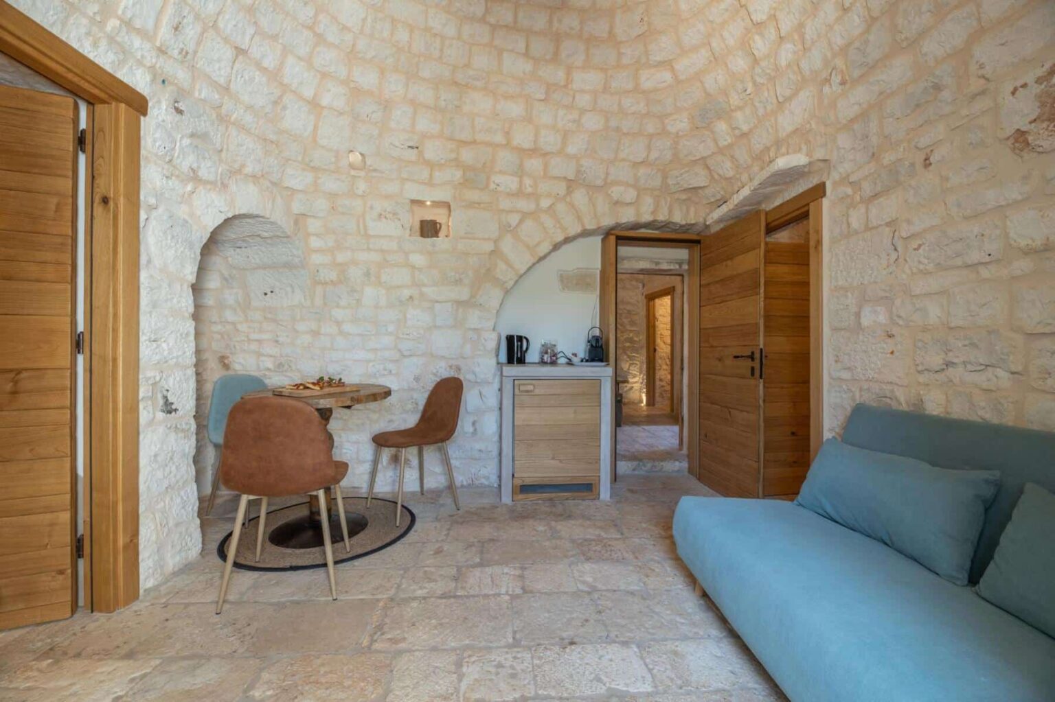 Cozy interior of a stone cottage featuring a circular design, wooden doors, a small dining area with chairs, and a blue sofa, showcasing rustic charm and modern comfort. | Only on SopranoVillas