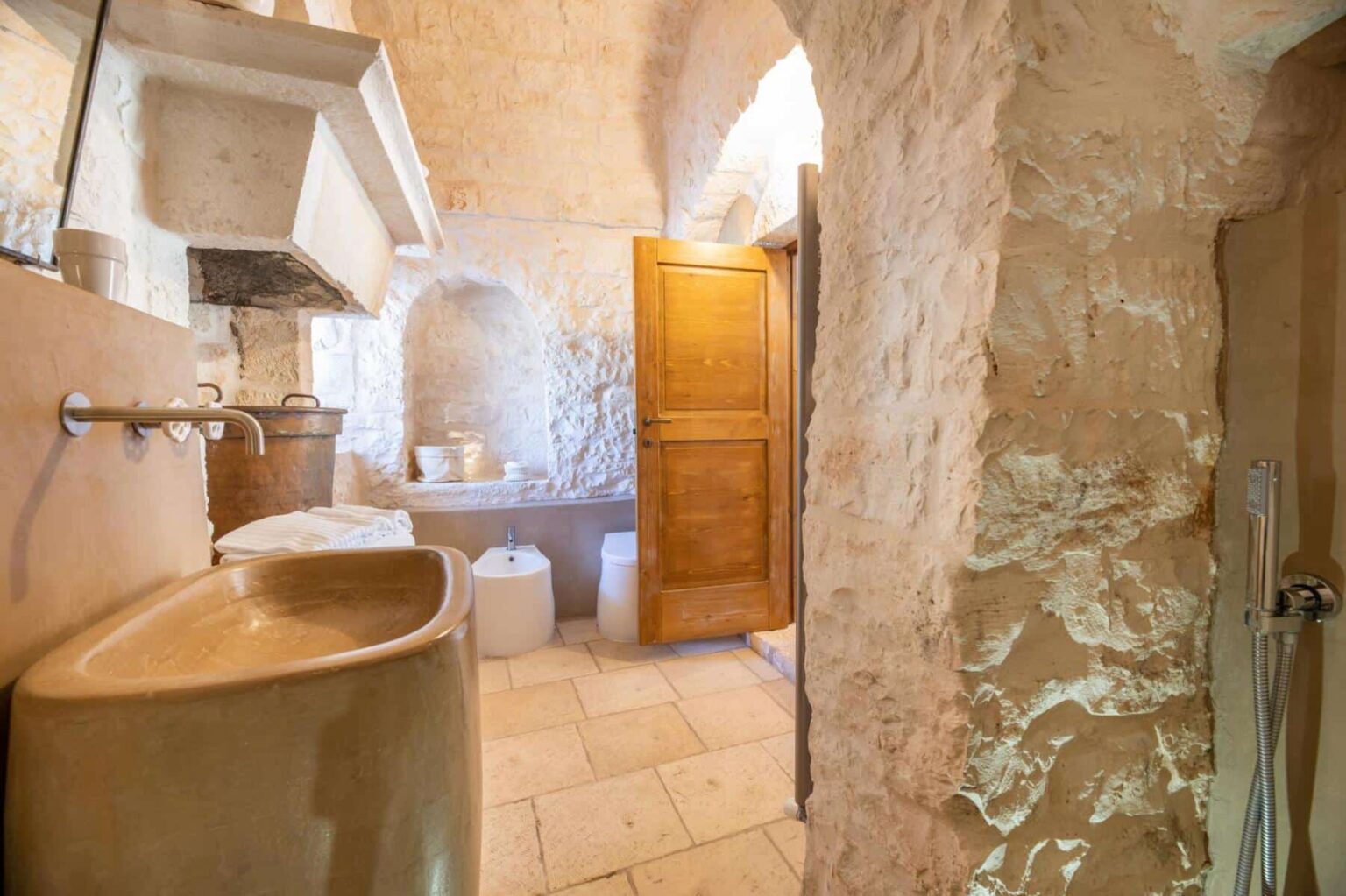Rustic bathroom interior featuring a stone wall, wooden door, and vintage sink. Natural light illuminates the space, highlighting the unique architectural details. | Only on SopranoVillas