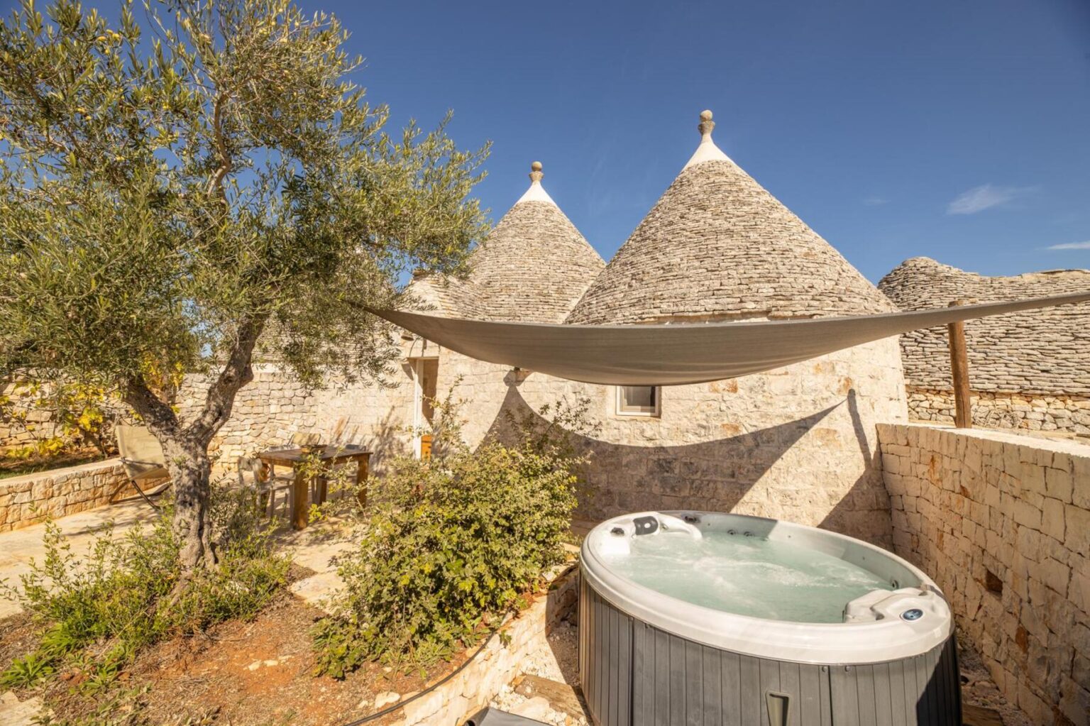 Tranquil outdoor space featuring a hot tub surrounded by lush greenery, traditional stone trulli houses, and a shaded seating area under a canopy, ideal for relaxation in a picturesque setting. | Only on SopranoVillas