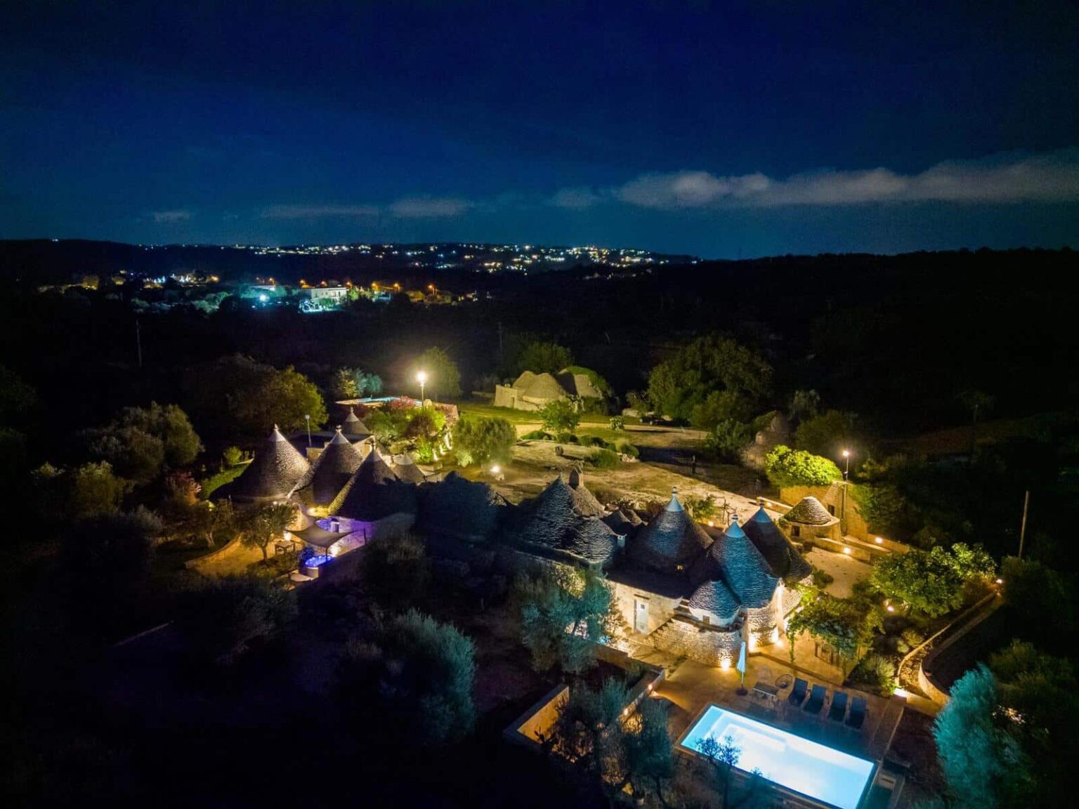 Aerial view of a beautifully lit rural property at night, featuring traditional thatched-roof cottages surrounded by greenery and a swimming pool, with distant city lights in the background. | Only on SopranoVillas
