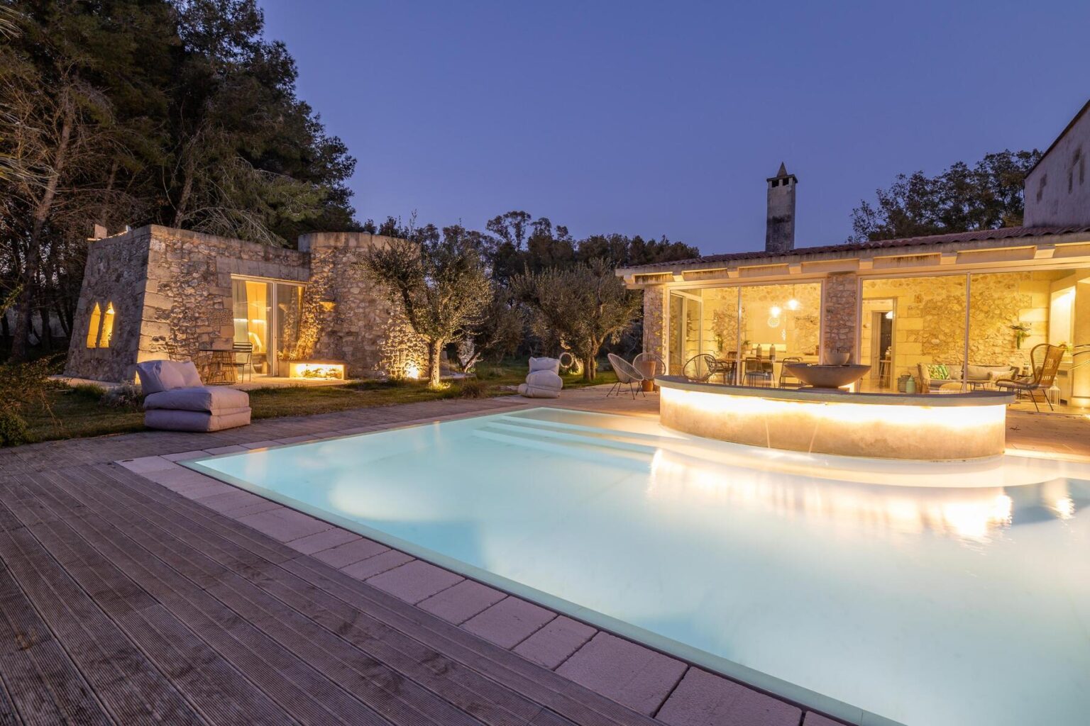 Luxurious outdoor pool area at dusk with illuminated stone building, modern bar, and cozy seating, surrounded by lush greenery. | Only on SopranoVillas