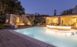 Luxurious outdoor pool area at dusk with illuminated stone building, modern bar, and cozy seating, surrounded by lush greenery.