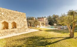 Stone villa with a lush green lawn and swimming pool, surrounded by olive trees under a clear blue sky. Ideal for a tranquil getaway or vacation rental.