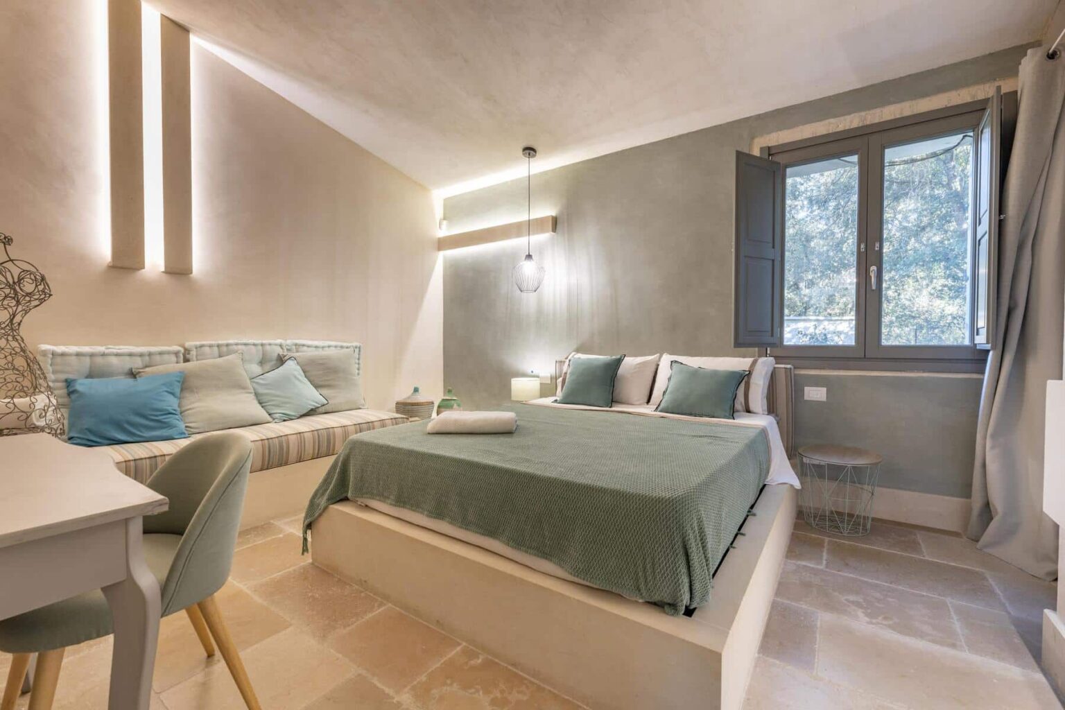 Cozy bedroom with a green bedspread, accent pillows, and a comfortable seating area. Features include soft lighting, a modern desk chair, and a window overlooking nature, creating a serene atmosphere ideal for relaxation. | Only on SopranoVillas