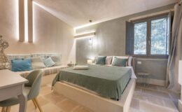 Cozy bedroom with a green bedspread, accent pillows, and a comfortable seating area. Features include soft lighting, a modern desk chair, and a window overlooking nature, creating a serene atmosphere ideal for relaxation.