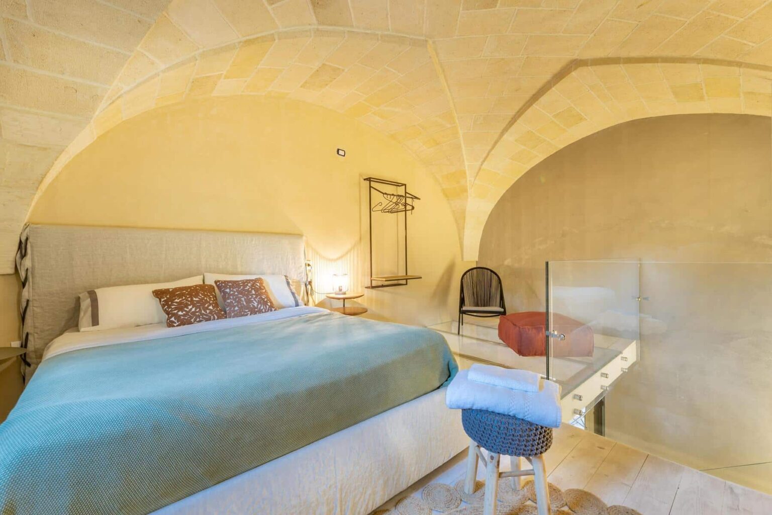 Cozy bedroom interior featuring a queen-sized bed with a blue throw blanket, minimalist decor, and an arched ceiling. The room includes a wooden chair, a glass partition, and warm lighting, creating a serene atmosphere ideal for relaxation. | Only on SopranoVillas