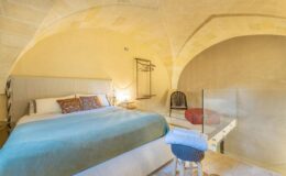 Cozy bedroom interior featuring a queen-sized bed with a blue throw blanket, minimalist decor, and an arched ceiling. The room includes a wooden chair, a glass partition, and warm lighting, creating a serene atmosphere ideal for relaxation.