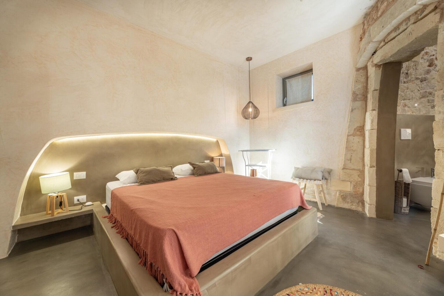 Modern hotel room featuring a cozy king-sized bed with a coral blanket, stylish bedside lamps, and minimalist decor. The room has a warm, neutral color palette and natural stone accents, creating a serene atmosphere. | Only on SopranoVillas