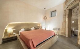 Modern hotel room featuring a cozy king-sized bed with a coral blanket, stylish bedside lamps, and minimalist decor. The room has a warm, neutral color palette and natural stone accents, creating a serene atmosphere.