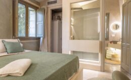 Cozy bedroom featuring a green bedspread, large windows with natural light, and a modern bathroom with glass enclosure. The interior design combines elegance and comfort, ideal for relaxation.