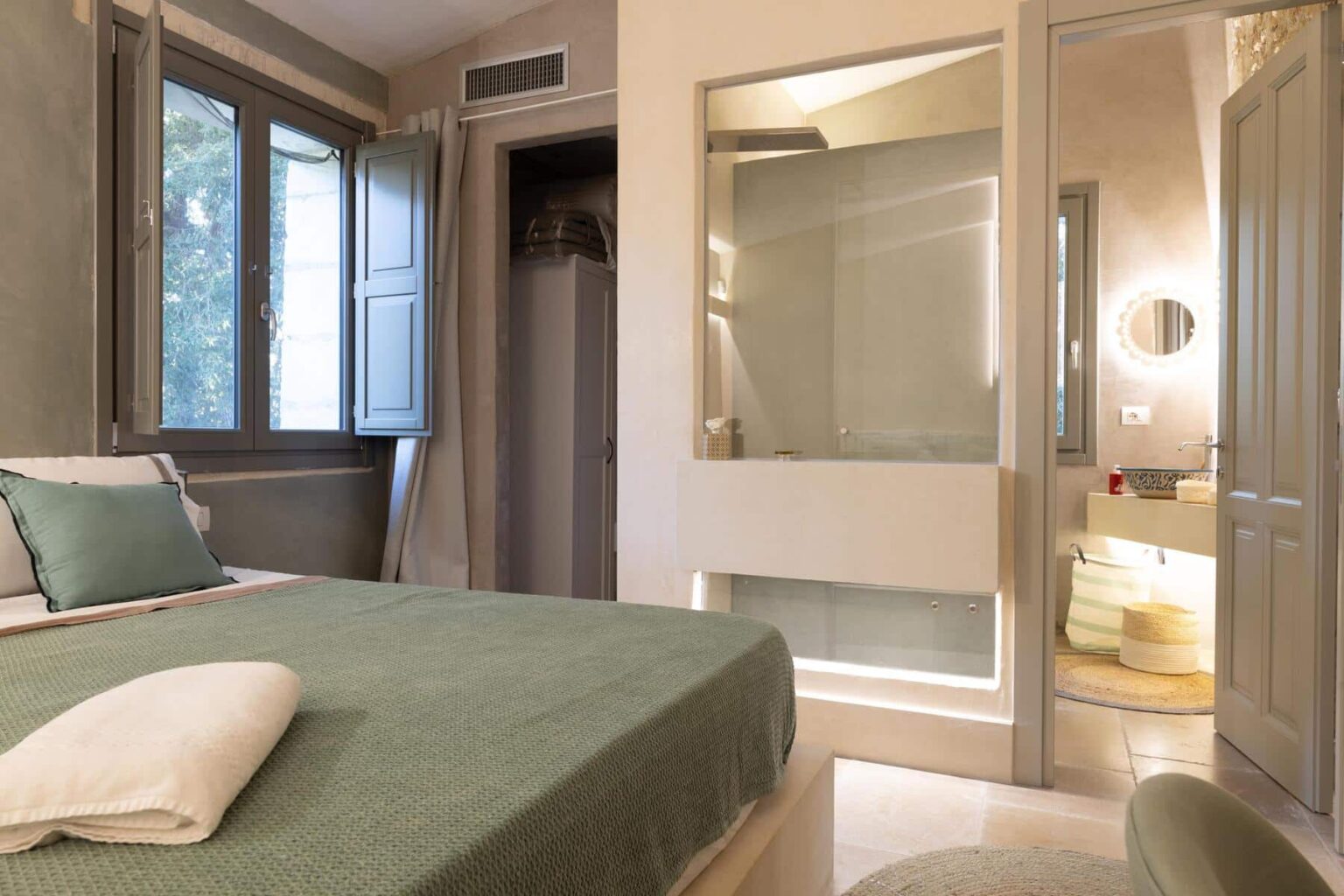 Modern bedroom interior featuring a green bedspread, natural light from large windows, and a stylish en-suite bathroom. The decor combines minimalism with comfort, ideal for a relaxing stay. | Only on SopranoVillas