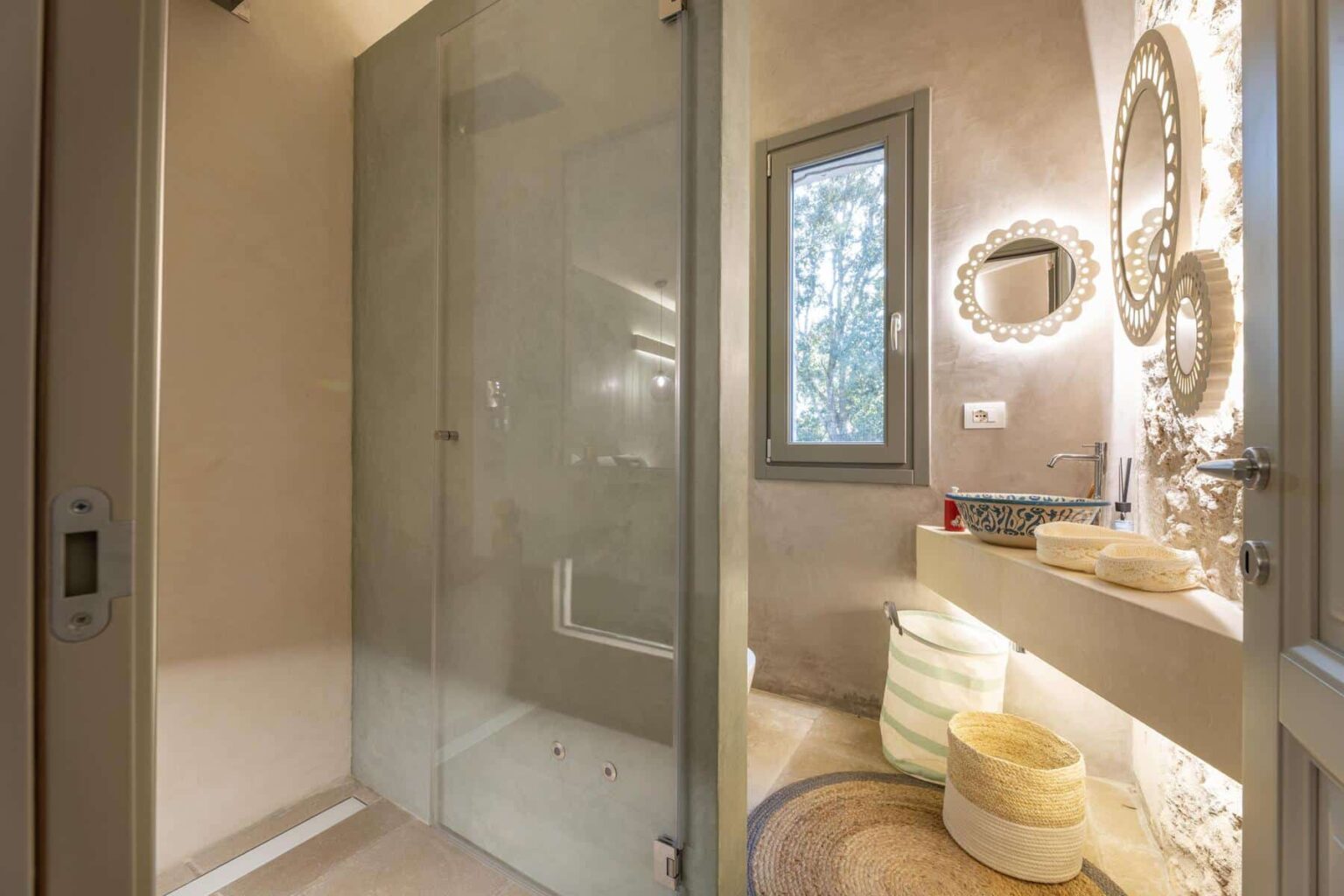 Modern bathroom featuring a glass shower enclosure, illuminated mirrors, and stylish decor. The design highlights neutral tones and elegant storage solutions, creating a serene and functional space. | Only on SopranoVillas