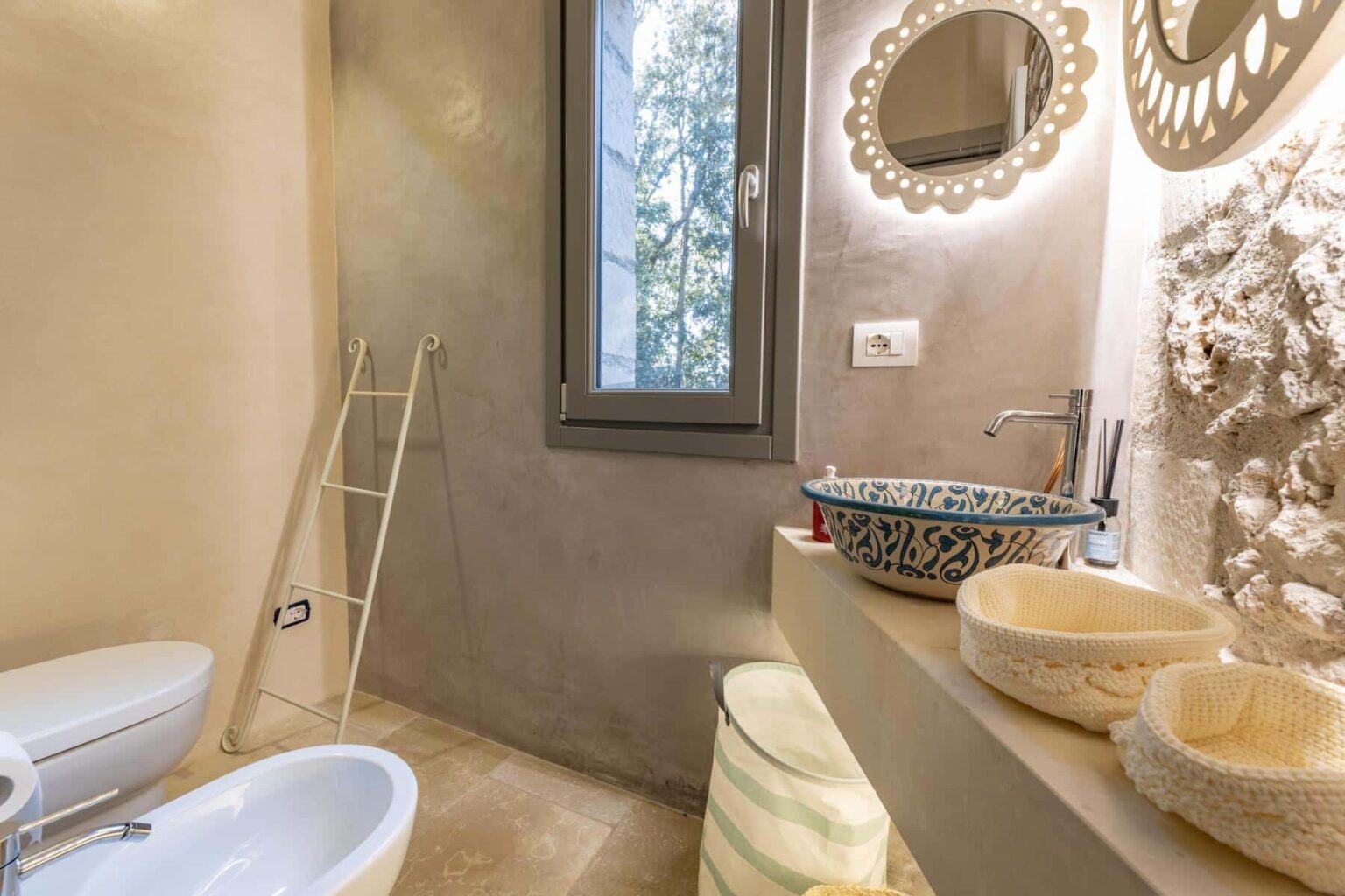 Modern bathroom featuring a minimalist design with a light color palette, stone accents, a stylish mirror, and decorative sink bowls. Natural light streams in through a window, enhancing the serene atmosphere. | Only on SopranoVillas