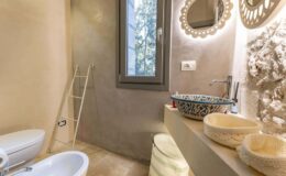 Modern bathroom featuring a minimalist design with a light color palette, stone accents, a stylish mirror, and decorative sink bowls. Natural light streams in through a window, enhancing the serene atmosphere.
