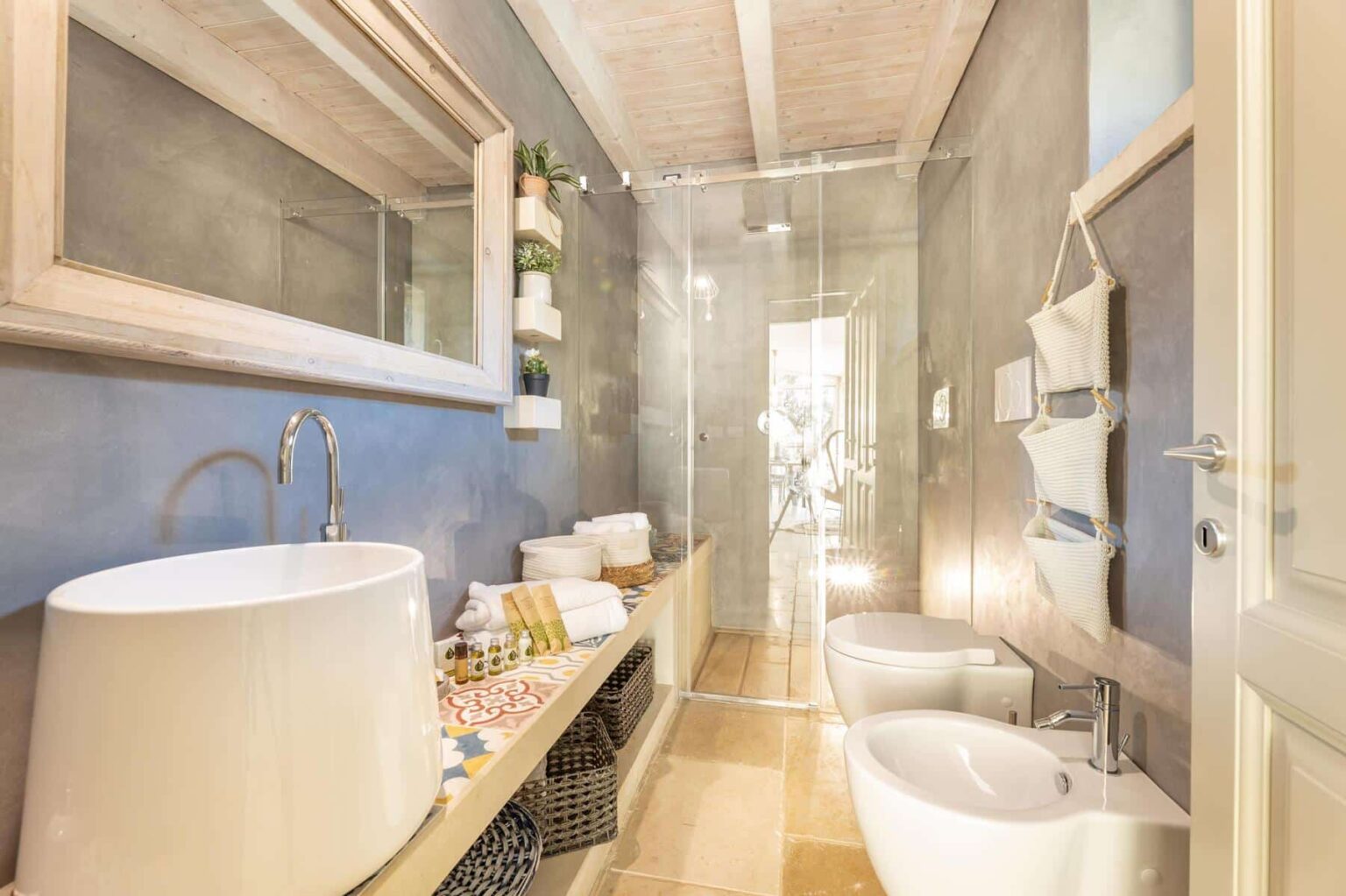 Modern bathroom interior featuring a stylish freestanding sink, glass shower enclosure, and elegant decor. The space includes a large mirror, neatly arranged towels, and decorative plants on wall shelves, creating a serene and functional atmosphere. | Only on SopranoVillas