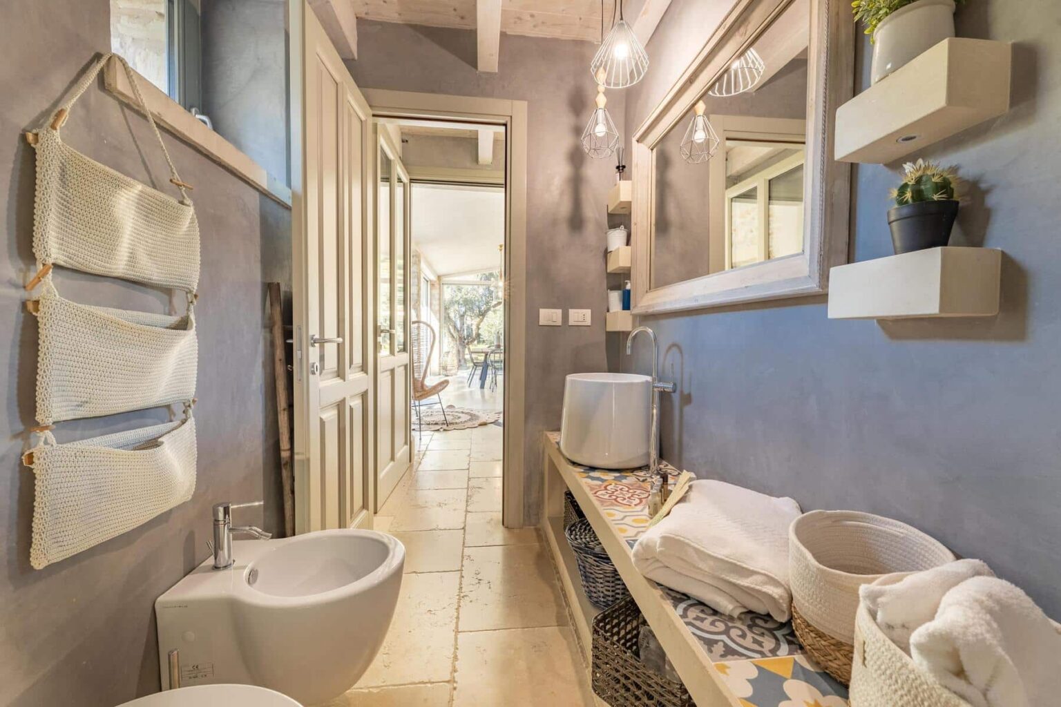 Modern bathroom interior featuring a stylish bidet, a sleek sink, and decorative storage baskets. The walls are painted in a soft gray, complemented by wooden accents and unique lighting fixtures. A large mirror reflects the natural light coming from the adjacent room, enhancing the spacious feel. | Only on SopranoVillas
