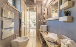 Modern bathroom interior featuring a stylish bidet, a sleek sink, and decorative storage baskets. The walls are painted in a soft gray, complemented by wooden accents and unique lighting fixtures. A large mirror reflects the natural light coming from the adjacent room, enhancing the spacious feel.