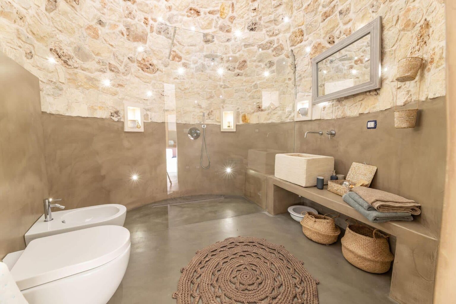 Modern bathroom interior featuring a curved stone wall, minimalist design, and natural materials. The space includes a white toilet, a walk-in shower with a sleek faucet, a unique stone sink, and a wooden bench with decorative baskets. Ambient lighting enhances the cozy atmosphere, while a round woven rug adds texture to the floor. | Only on SopranoVillas
