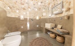 Modern bathroom interior featuring a curved stone wall, minimalist design, and natural materials. The space includes a white toilet, a walk-in shower with a sleek faucet, a unique stone sink, and a wooden bench with decorative baskets. Ambient lighting enhances the cozy atmosphere, while a round woven rug adds texture to the floor.