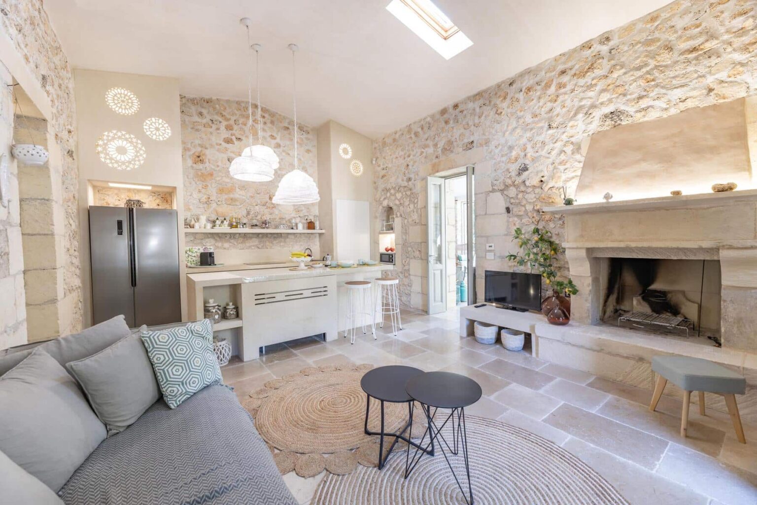 Modern living room with stone walls, featuring a cozy seating area, a stylish coffee table, a functional kitchen with a refrigerator, and a fireplace, illuminated by natural light. | Only on SopranoVillas