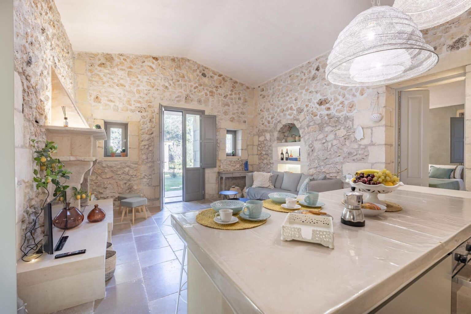 Cozy kitchen and living area featuring rustic stone walls, modern lighting, and a view of the garden through open doors. A dining table is set with cups, fruits, and a coffee maker, creating an inviting atmosphere. | Only on SopranoVillas