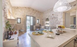Cozy kitchen and living area featuring rustic stone walls, modern lighting, and a view of the garden through open doors. A dining table is set with cups, fruits, and a coffee maker, creating an inviting atmosphere.