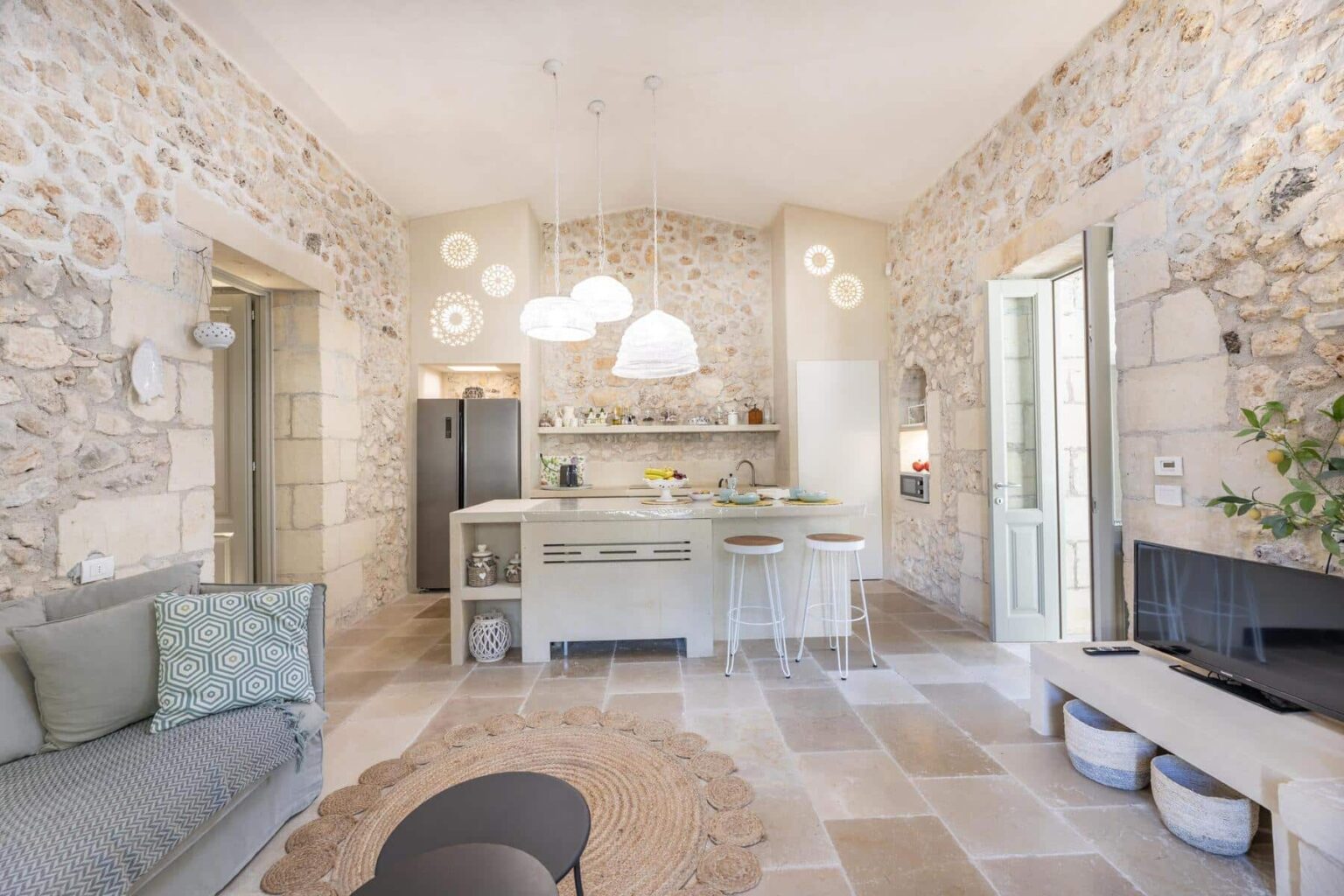 Modern open-concept kitchen and living area featuring natural stone walls, contemporary furniture, and stylish pendant lighting. The kitchen includes a central island with seating, sleek appliances, and decorative accents, while a cozy seating area showcases a patterned cushion and a round rug. | Only on SopranoVillas